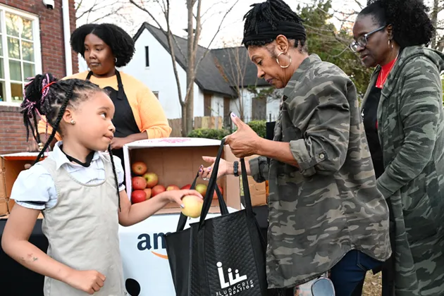 A woman and a young girl select apples from a stand, placing them in a black grocery bag.